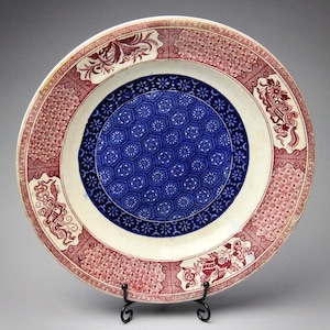 Peut inclure: Une assiette antique décorative avec un design détaillé. L'assiette présente un centre bleu et blanc avec un motif floral, entouré d'une bande blanche et d'une bordure rouge à motifs. Des motifs floraux et géométriques rouges ornés sont visibles.
