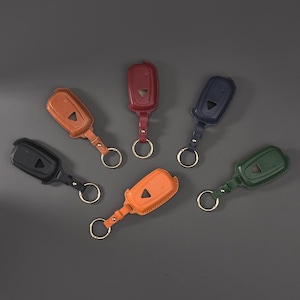 May include: Six leather car key fob covers in black, tan, orange, dark red, navy blue, and forest green. Each cover features a triangular cutout, visible stitching along the edges, a short leather strap, and a circular metal split ring for keys.