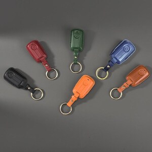May include: Six rectangular key fob covers in black, red, dark green, blue, orange, and tan. Each cover features stitched edges, a matching leather strap, and a circular gold-toned metal key ring attached to the base for securing keys.