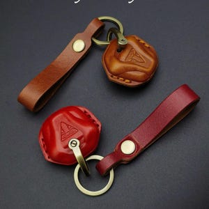 May include: Two leather motorcycle key covers with the text Motorcycle key cover. One cover is tan and the other is red. Each features a brass-toned metal ring, a matching snap button, and a molded leather pouch embossed with a Triumph logo.