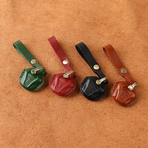 May include: Four leather key fob covers in green, red, black, and brown. Each cover features a molded geometric shape, a matching leather strap with a brass-toned snap button, and a brass-toned metal ring attachment for securing keys.