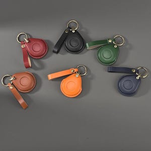 Op de afbeelding: Zes leren sleutelhangers in rood, zwart, groen, bruin, oranje en marineblauw. Elke sleutelhanger heeft een ronde houder met een bijpassende leren riem, een metalen ring en een kleine metalen klinknagel. De houders hebben een gladde, ronde vorm.