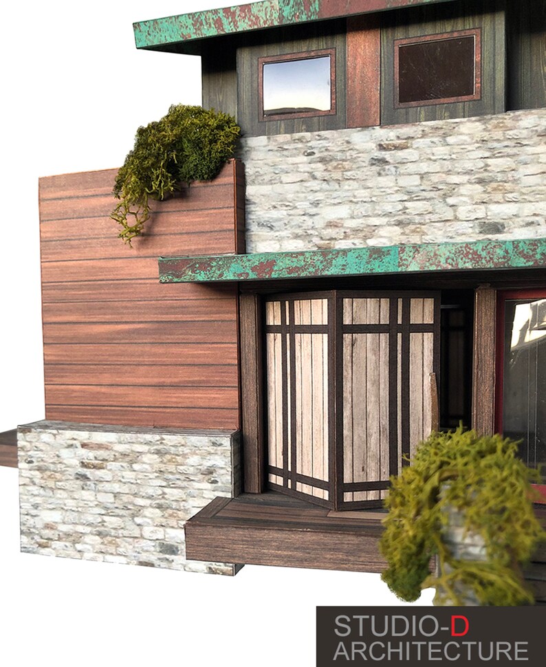 K&ouml;nnte beinhalten: Ein Modellhaus mit einer Fassade aus Stein und Holz. Das Haus hat ein gro&szlig;es Fenster und eine T&uuml;r mit einem Holzrahmen. Das Haus ist von Gr&uuml;n umgeben.