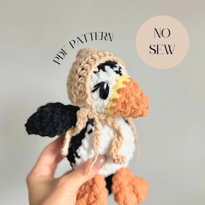 May include: A handmade crochet puffin bird with a tan hat and orange beak and feet. The bird is primarily white with black wings. The image includes the text "PDF PATTERN" and "NO SEW". The store name "Klein Kreations Store" is also visible.