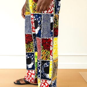 May include: Colorful patchwork pants with a vibrant mix of patterns and colors, including red, yellow, blue, and black. The pants are paired with black sandals. The design features geometric shapes and abstract motifs.
