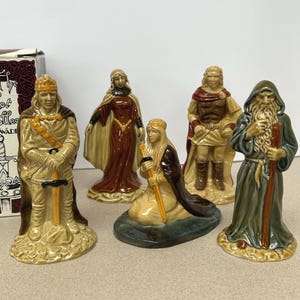 May include: A collection of five ceramic figurines from the Camelot Collection. The figurines depict characters in various poses, including a king, a kneeling figure, and a wizard. The figures are painted in tan, brown, and green, with gold accents. A box is visible.