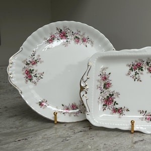 May include: A two-piece porcelain serving set. A round plate and a rectangular tray, both white with a floral pattern of pink roses and green leaves. Gold trim accents the edges and handles. The set is displayed on gold stands.