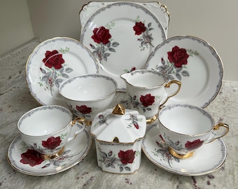 May include: A white porcelain tea set featuring red rose patterns, gray foliage, and gold trim. The set includes a square serving plate, two round plates, two teacups with saucers, a creamer pitcher, a sugar bowl with a lid, and a small bowl.