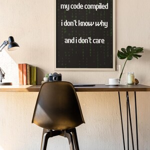 Pu&ograve; includere: Una stampa incorniciata con sfondo nero e testo bianco che recita "my code compiled i don't know why and i don't care". Il testo &egrave; circondato da una matrice verde di numeri.