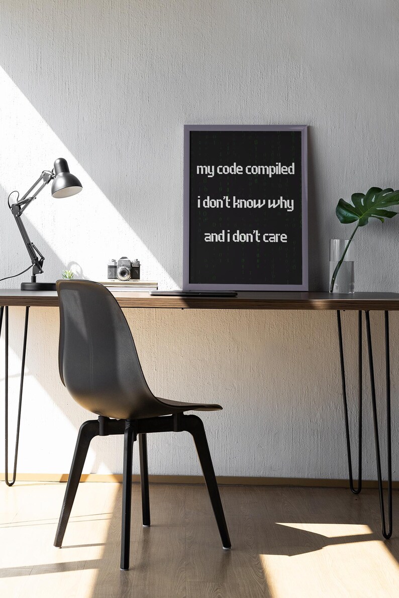Pu&ograve; includere: Un cartello nero incorniciato con testo bianco che recita "my code compiled i don't know why and i don't care". Il cartello &egrave; su una scrivania di legno con una sedia nera, una lampada, una pianta in vaso e una macchina fotografica.