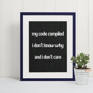 Pu&ograve; includere: Una stampa incorniciata con sfondo nero e testo verde che recita "my code compiled i don't know why and i don't care". La stampa &egrave; in una cornice blu scuro e si trova su una superficie bianca.