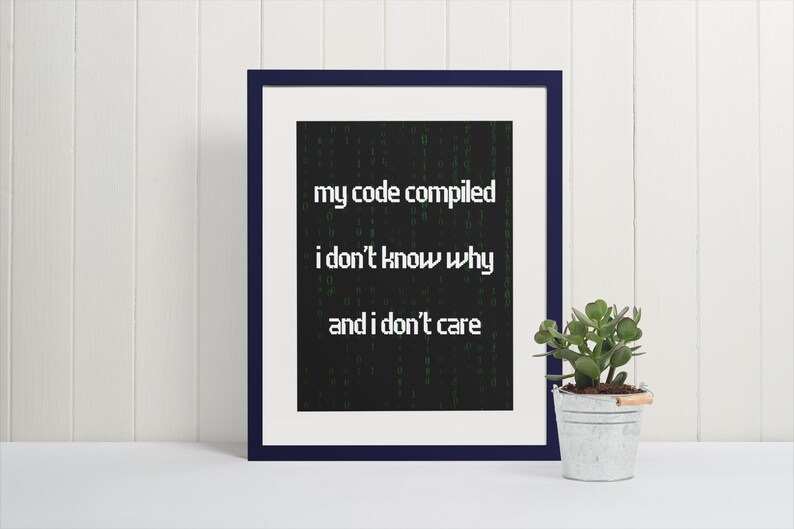 Pu&ograve; includere: Una stampa incorniciata con sfondo nero e testo verde che recita "my code compiled i don't know why and i don't care". La stampa &egrave; in una cornice blu scuro e si trova su una superficie bianca.