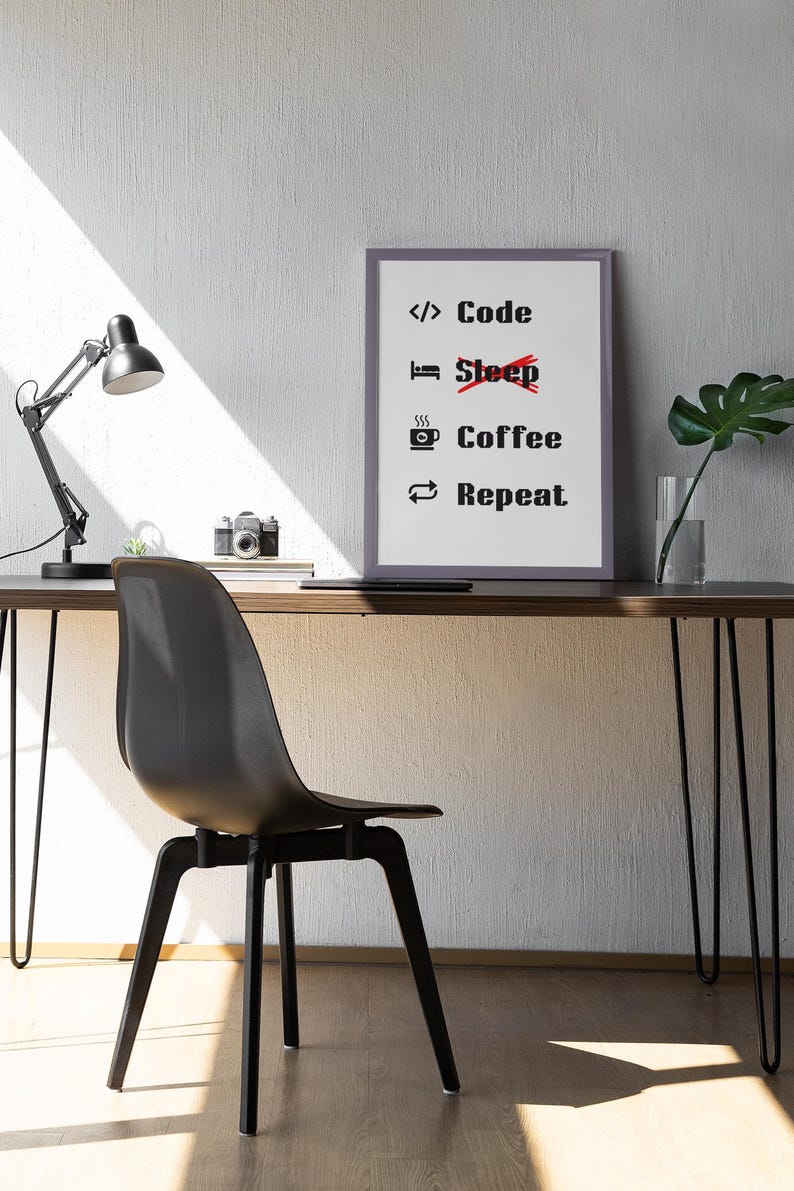 Pode incluir: Uma impress&atilde;o emoldurada com o texto "Code Coffee Repeat" com uma linha vermelha atravessando a palavra "Sleep". A impress&atilde;o est&aacute; pendurada em uma parede branca acima de uma mesa com uma cadeira preta, uma l&acirc;mpada e uma planta em vaso.