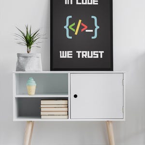 Puede incluir: Un armario blanco con una puerta negra y dos estantes. El estante superior tiene un cono de helado azul y blanco. El estante inferior tiene una pila de libros. Un p&oacute;ster enmarcado negro con el texto "In code we trust" y un s&iacute;mbolo de corchete de c&oacute;digo colorido est&aacute; colgado en la pared sobre el armario.