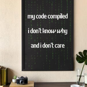 Pu&ograve; includere: Una stampa incorniciata nera con sfondo bianco e testo verde che recita "my code compiled i don't know why and i don't care". Il testo &egrave; circondato da uno schema di 0 e 1 verdi.