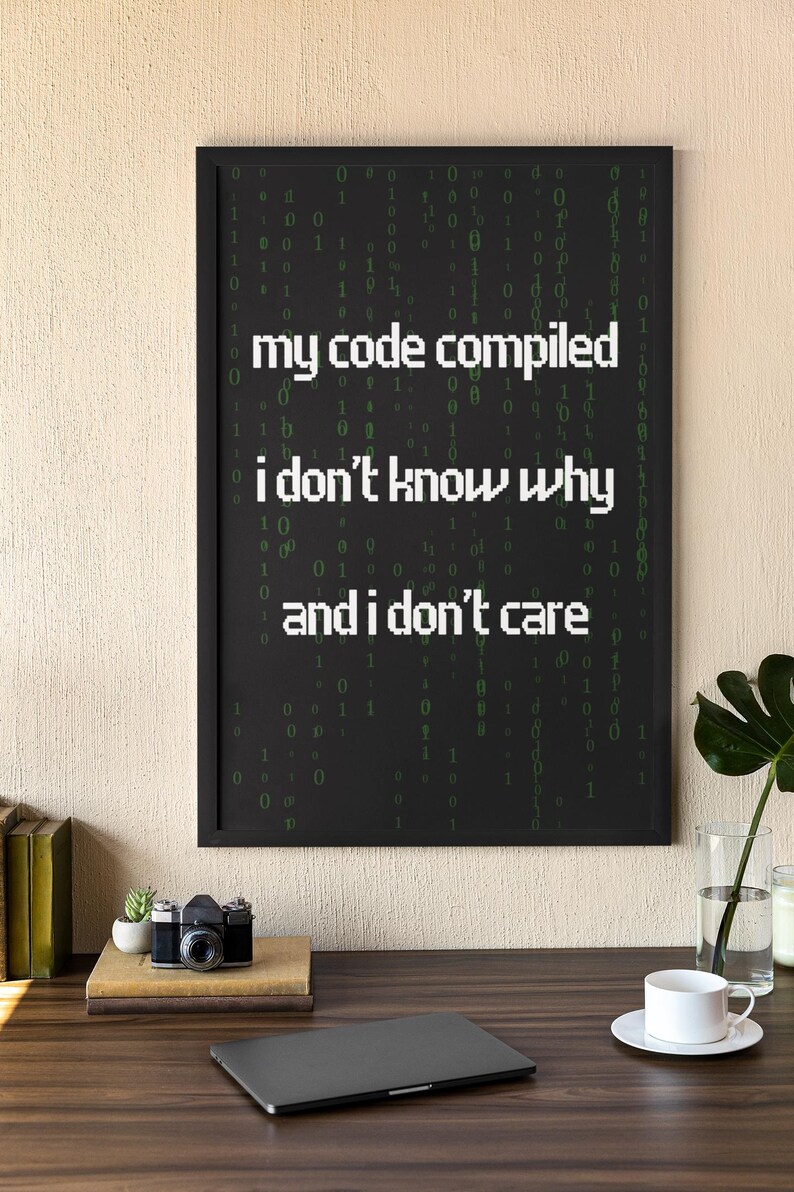 Pu&ograve; includere: Una stampa incorniciata nera con sfondo bianco e testo verde che recita "my code compiled i don't know why and i don't care". Il testo &egrave; circondato da uno schema di 0 e 1 verdi.