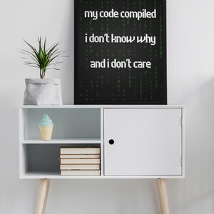 Pu&ograve; includere: Un poster incorniciato nero con testo verde che recita "my code compiled i don't know why and i don't care" su una parete bianca sopra un armadio bianco con una porta chiusa e due ripiani. Lo scaffale superiore ha un cono gelato blu e bianco e lo scaffale inferiore ha una pila di libri. L'armadio &egrave; su un tappeto a motivi bianco e nero.