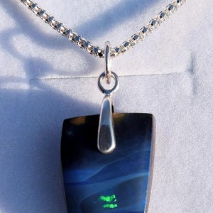 May include: A rectangular pendant with a silver bail hangs from a silver chain. The pendant features a dark blue and black stone with a flash of green. The chain is a delicate, woven design.