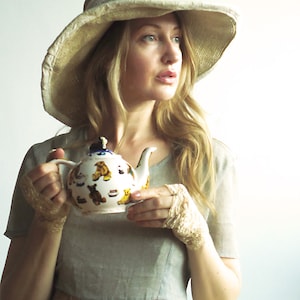May include: A woman wearing a wide-brimmed straw hat and a light gray dress holds a white teapot with a floral pattern. The teapot has a blue handle and a gold lid.