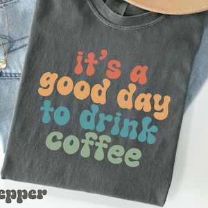 Könnte beinhalten: Ein dunkelgraues T-Shirt mit dem Schriftzug "it's a good day to drink coffee" in orange, blau und grün. Das Shirt wird mit hellblauen Jeans und einem beigefarbenen Hut kombiniert. Das Wort "Pepper" steht unten.