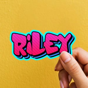 May include: A sticker with the name "Riley" in a graffiti-style font. The letters are pink with a striped pattern, outlined in blue and black, and set against a yellow background.