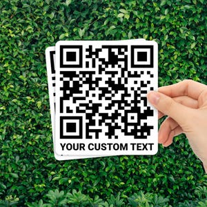 May include: Two white square QR code stickers with black and white patterns and the text "YOUR CUSTOM TEXT" are held against a background of green leaves. The QR codes are a modern design element.