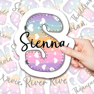 May include: A sticker featuring the name "Sienna" in a black script font, overlaid on a large, pastel-colored letter "S". The letter is decorated with white hearts and a stitched border. Other stickers with names are in the background.