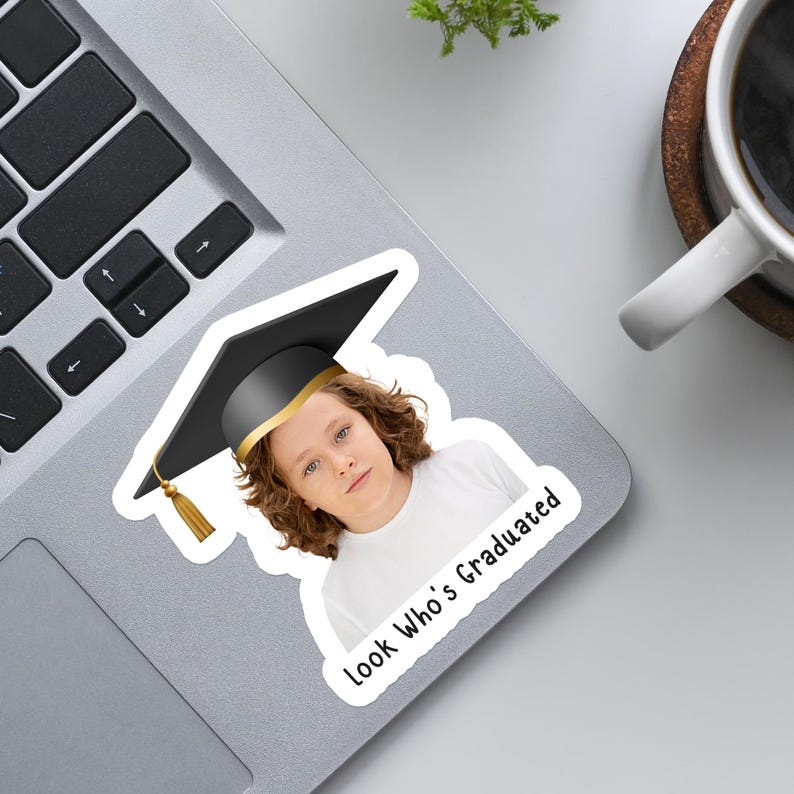 May include: A sticker featuring a person wearing a graduation cap with a gold band and tassel. The sticker has the text "Look Who's Graduated" in a black, handwritten font.