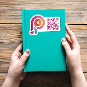 May include: A teal-colored book with a white sticker featuring a stylized pink and orange logo and a QR code. The book is held by two hands with manicured nails, set against a wooden background.