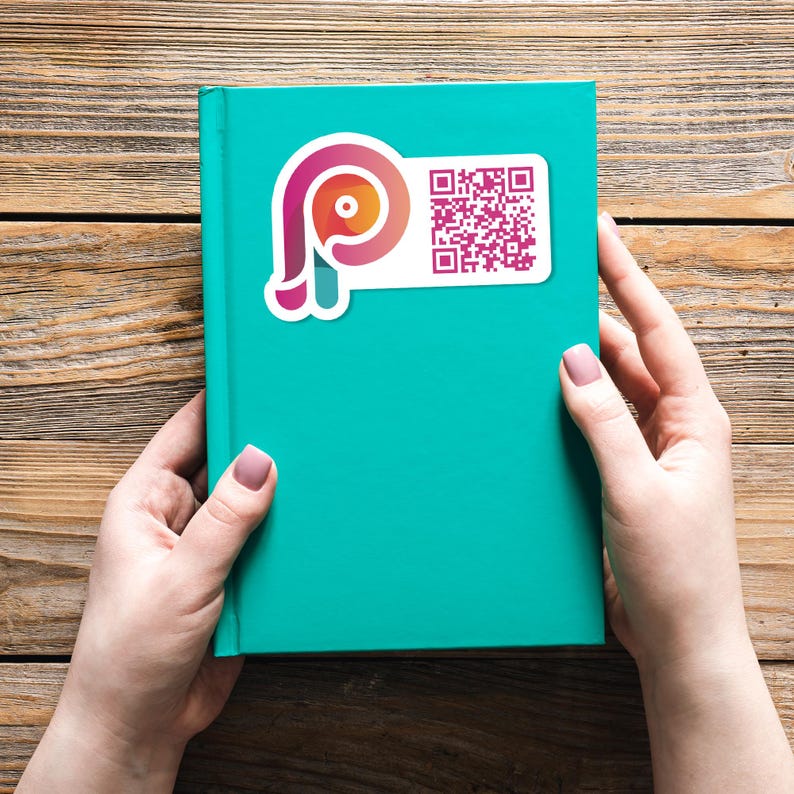 May include: A teal-colored book with a white sticker featuring a stylized pink and orange logo and a QR code. The book is held by two hands with manicured nails, set against a wooden background.
