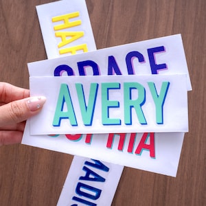 May include: A stack of rectangular white stickers featuring names in bold, colorful, block-style lettering with drop shadows. The top sticker displays the name AVERY in mint green, while others show names like HAP, SPACE, and SOPHIA in yellow, blue, and red.