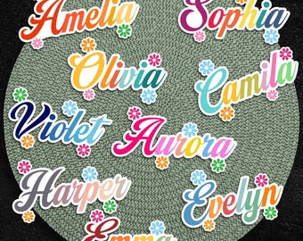 May include: A collection of colorful name stickers, including "Amelia," "Sophia," "Olivia," and others, each with a unique font and floral accents. The stickers are arranged on a textured, circular background.