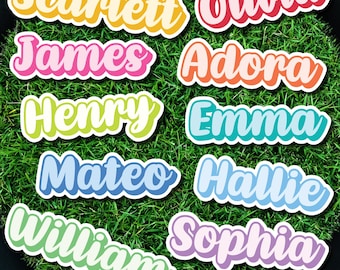 May include: Colorful name stickers with names like Scarlett, Olivia, James, Adora, Henry, Emma, Mateo, Hallie, William, and Sophia. Each sticker has a white border and is set against a green, grassy background.