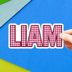 May include: A die-cut sticker featuring the name LIAM in bold, block capital letters. The letters have a red and white gingham checkered pattern with a dark red outline and a thick white border surrounding the entire name design.