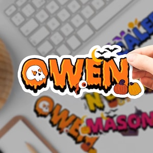 May include: A die-cut vinyl sticker featuring the name OWEN in a dripping orange font with a black outline. The design includes a white skull with fire in its eye sockets, a yellow crescent moon, black bats, a single eyeball, and two pumpkins.