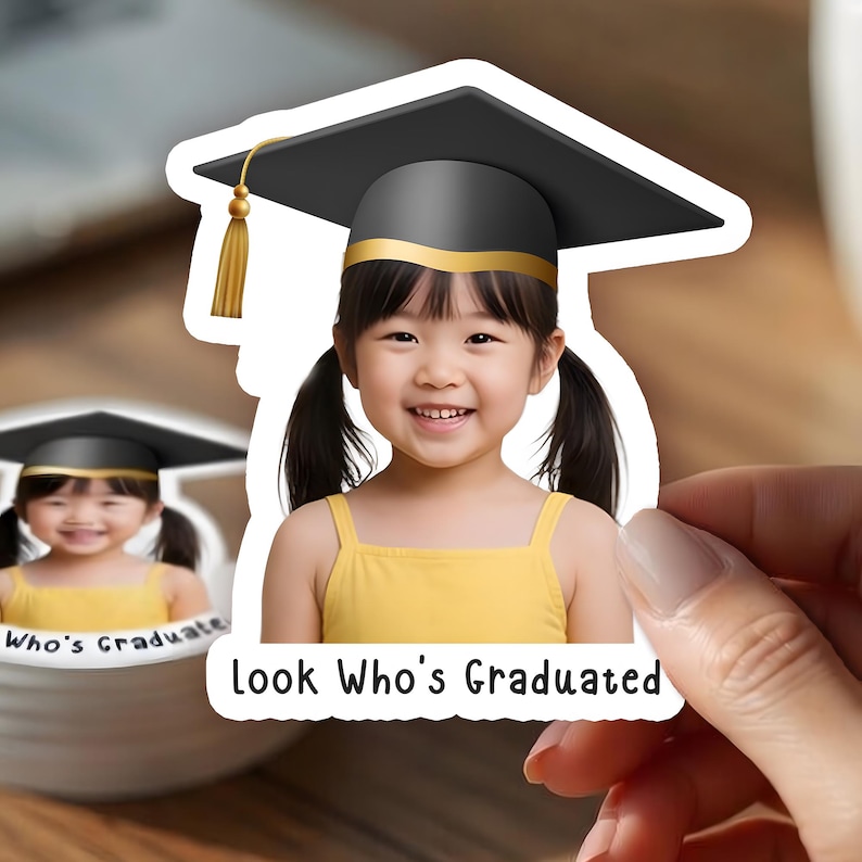 May include: A sticker featuring a smiling child wearing a graduation cap. The cap is black with a gold tassel and trim. The child is wearing a yellow top. The text "Look Who's Graduated" is below the image.