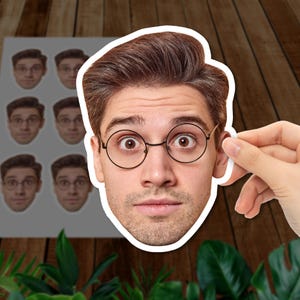 May include: A sheet of stickers featuring a man's face with round glasses and a surprised expression. The stickers are white with a detailed image of the man's face. The background includes a wooden surface and green plants.