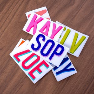 May include: A collection of rectangular white stickers featuring bold, colorful block letters. The stickers display names and words including KAY, SOP, ZOE, and IV in vibrant shades of pink, blue, green, and orange with contrasting outlines.