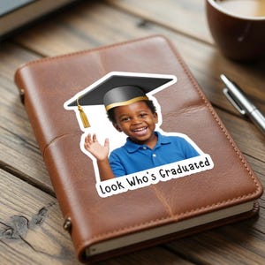 May include: A sticker featuring a smiling child wearing a graduation cap. The child is wearing a blue polo shirt. The text "Look Who's Graduated" is below the child. The sticker is on a brown leather notebook.
