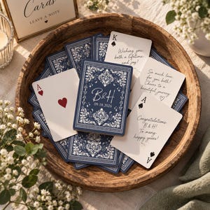 May include: A collection of custom playing cards in a wooden bowl. The cards feature a dark blue back with white ornate patterns, the initials B&H, and the date 09.18.2026. Some cards show handwritten messages of congratulations and well wishes.