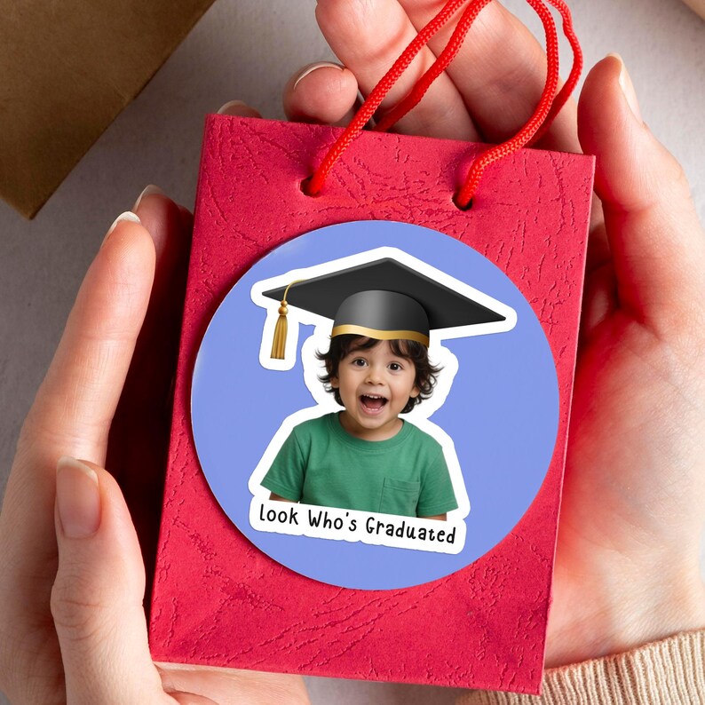 May include: A red gift bag with red handles is held by a person. A round sticker on the bag features a child wearing a graduation cap and the text "Look Who's Graduated". The child is wearing a green shirt and has a surprised expression.
