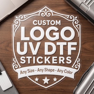 May include: A clear, shield-shaped sticker featuring white text that reads CUSTOM LOGO UV DTF STICKERS, with the additional text Any Size - Any Shape - Any Color on a banner, accented by three white stars at the bottom and decorative scrollwork.