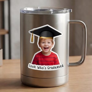 May include: Stainless steel travel mug with a sticker of a smiling child wearing a graduation cap. The child is wearing a red and black striped shirt. The sticker has the text "Look Who's Graduated".