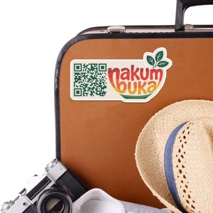 May include: A sticker with the text "nakum buka" in orange, yellow, and green, and a QR code, is affixed to a brown suitcase. A straw hat and a vintage camera are also visible.