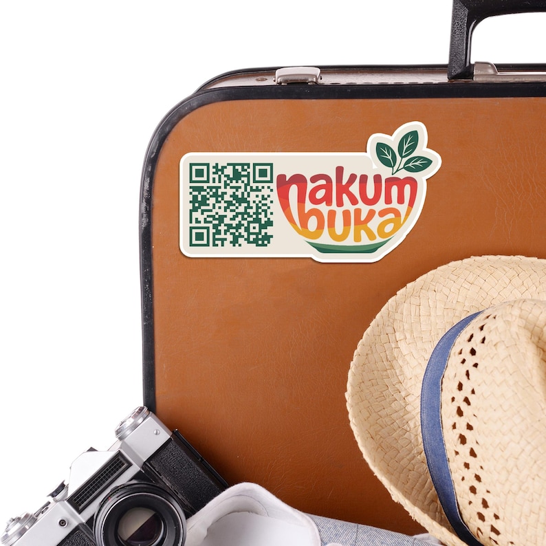 May include: A sticker with the text "nakum buka" in orange, yellow, and green, and a QR code, is affixed to a brown suitcase. A straw hat and a vintage camera are also visible.