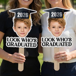 May include: Two graduation photo props featuring a child's face wearing a graduation cap with the year 2026. The props have the text "LOOK WHO'S GRADUATED" in bold, white letters. The props are held up by wooden sticks.