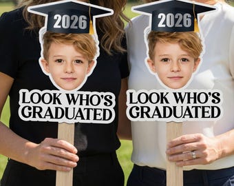 May include: Two graduation photo props featuring a child's face wearing a graduation cap with the year 2026. The props have the text "LOOK WHO'S GRADUATED" in bold, white letters. The props are held up by wooden sticks.