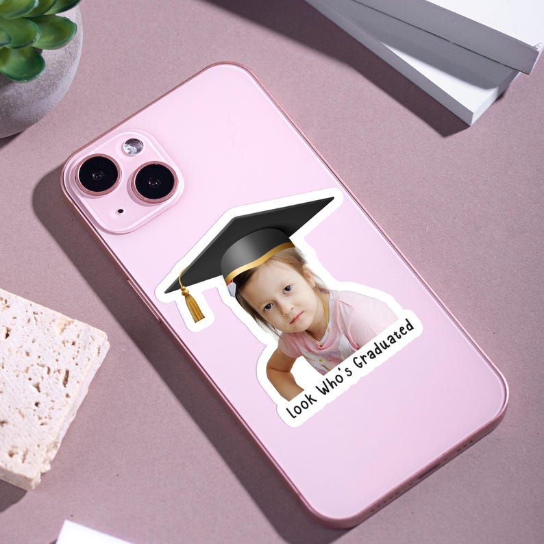 May include: A pink smartphone with a sticker featuring a child wearing a graduation cap. The sticker has the text "Look Who's Graduated". The cap is black with a gold band and tassel.