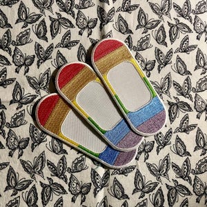 May include: Three rainbow-colored fabric patches with white centers. The patches are arranged in a row, with the top patch slightly overlapping the middle patch, and the middle patch slightly overlapping the bottom patch. The patches are on a white fabric background with a black butterfly pattern.