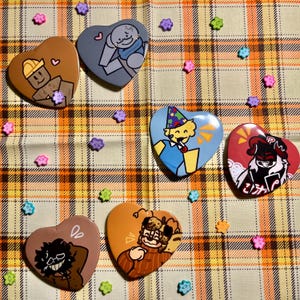 May include: Six heart-shaped pins with various cartoon characters. The pins are on a checkered orange and brown fabric background. The characters include a person wearing a blue hat, a person with a blue and white striped shirt, a person with a brown hat, a person with a red and black shirt, a person with a brown shirt, and a person with a black shirt.